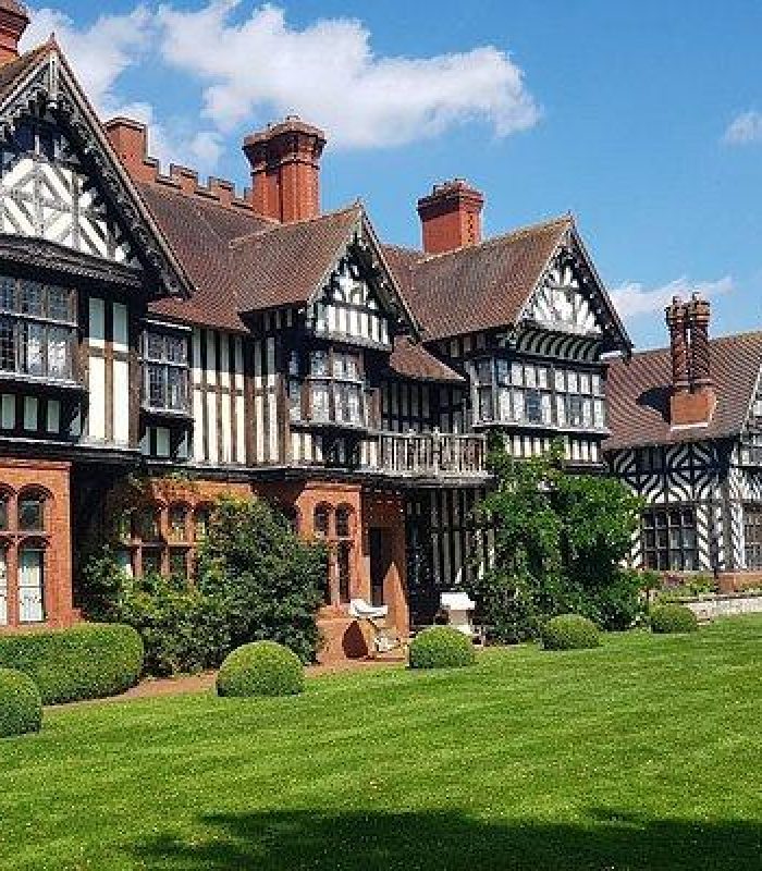 Un grand manoir historique de style Tudor avec une charpente en bois, de hautes cheminées et des pelouses vertes entretenues sous un ciel bleu brillant avec des nuages épars.