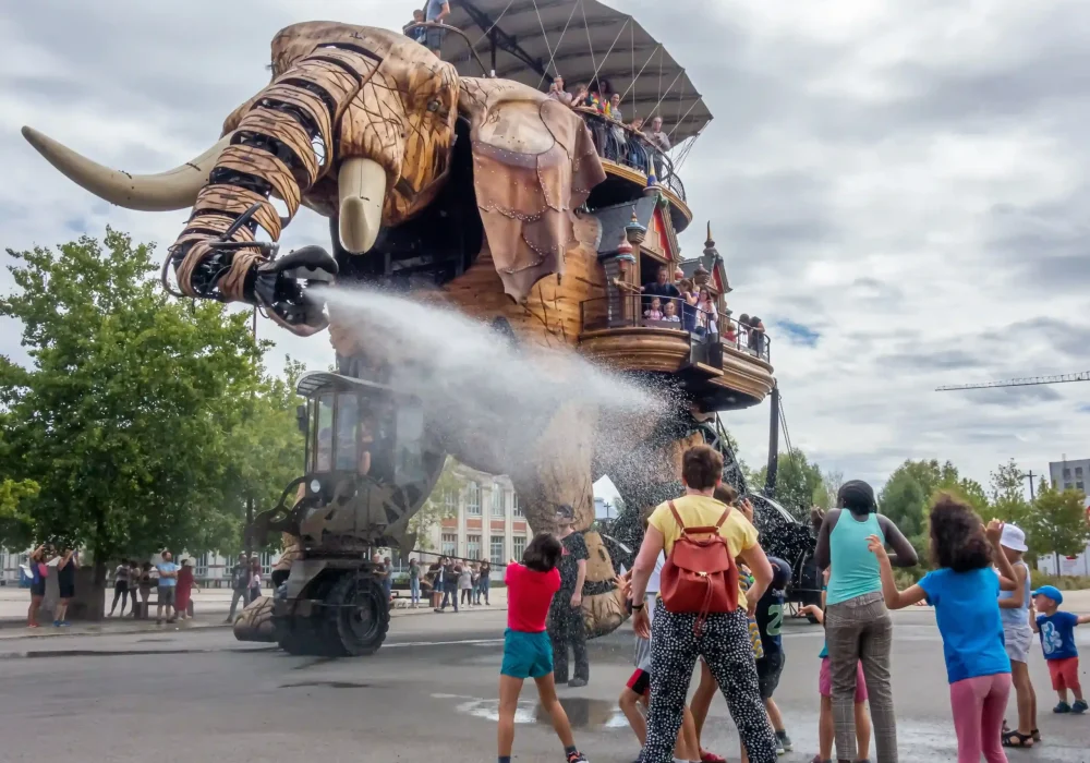 Un grand éléphant mécanique asperge d'eau un groupe d'enfants et d'adultes enthousiastes sur une place publique ; plusieurs personnes sont assises sur son dos, à l'abri d'un auvent. Le ciel est nuageux et des arbres se profilent à l'arrière-plan.