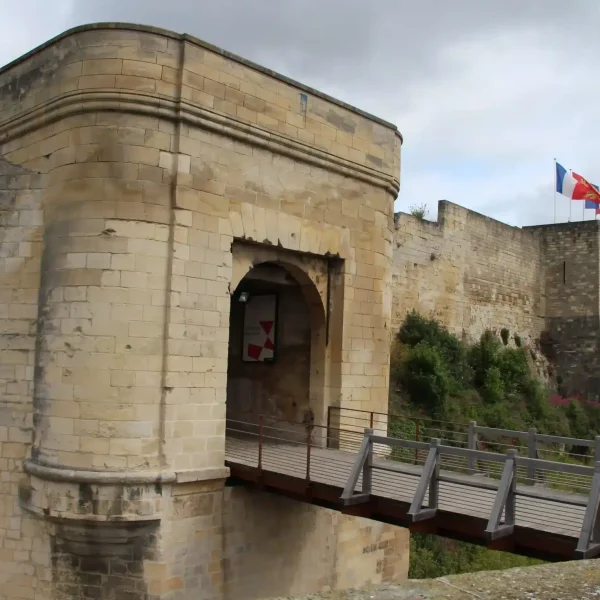 Une forteresse en pierre avec une entrée voûtée, un pont en bois et des drapeaux flottant au-dessus des remparts par temps nuageux.
