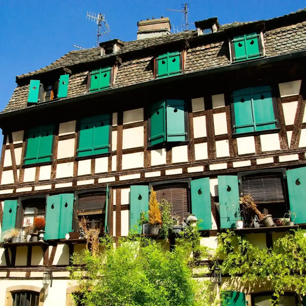Une maison traditionnelle à colombages, dotée de volets verts, se détache sous un ciel d'un bleu limpide ; on y remarque des plantes sur les rebords de fenêtre et des antennes de télévision qui dépassent du toit.
