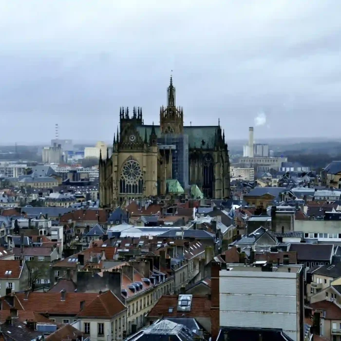 Vue aérienne d'une ville européenne aux bâtiments densément alignés, avec une grande cathédrale de style gothique aux hautes flèches et aux vitraux, sous un ciel nuageux.