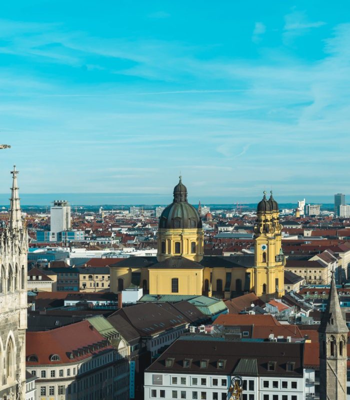 Vue aérienne du paysage urbain de Munich avec, à gauche, la tour de l'horloge du nouvel hôtel de ville et, à l'arrière-plan, l'église jaune des Théatins, des bâtiments aux toits rouges et un ciel bleu limpide.