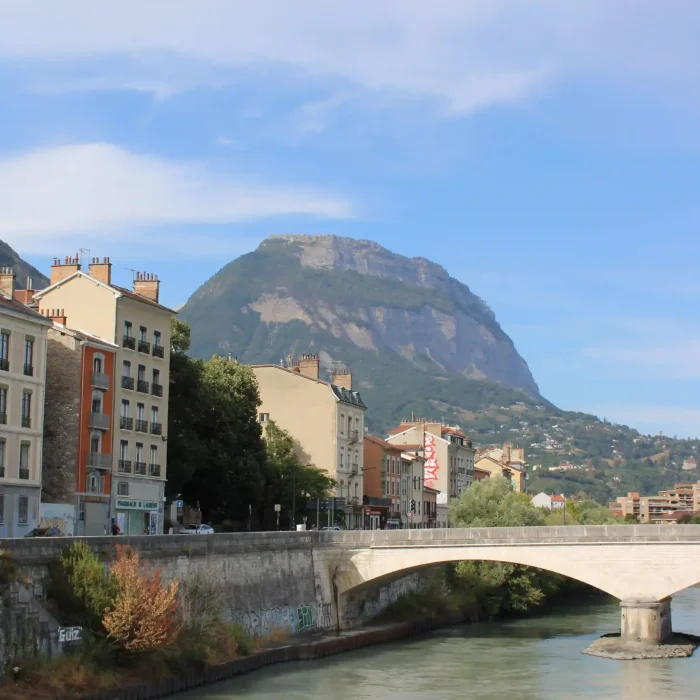 Un pont en pierre enjambe une rivière bordée de bâtiments colorés, avec en arrière-plan une grande montagne boisée qui se détache sous un ciel bleu parsemé de légers nuages.
