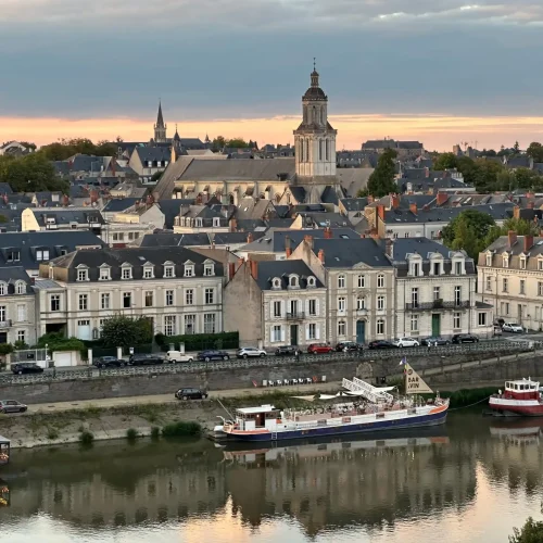 Au coucher du soleil, cette ville européenne au bord d'un fleuve offre une vue sur ses bâtiments historiques, le clocher d'une église et des bateaux amarrés le long de l'eau calme, qui reflète son architecture pittoresque.