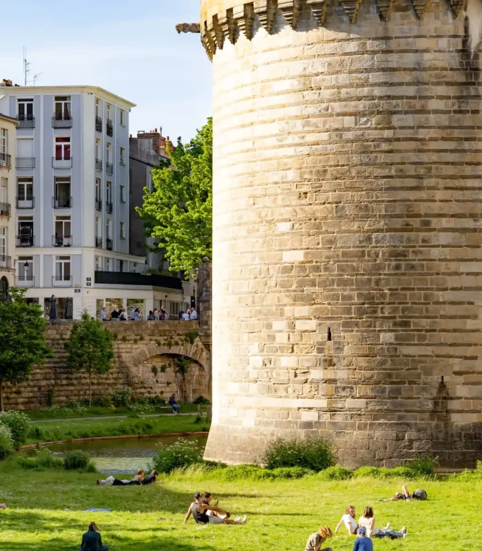 Par une journée ensoleillée, des gens se détendent sur une pelouse, près d'une grande tour en pierre, avec un immeuble d'habitation et des arbres en arrière-plan.