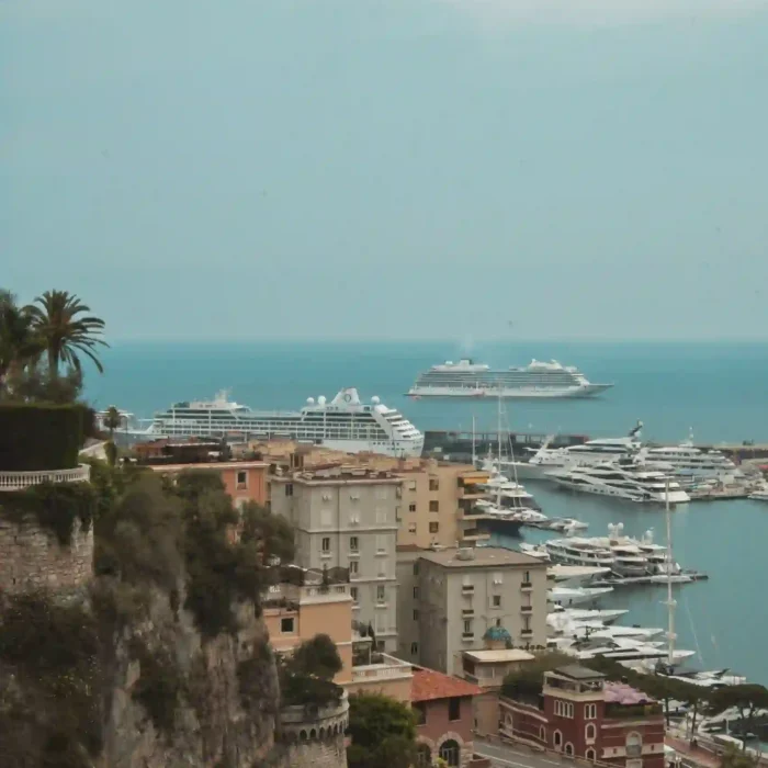 Vue d'un port méditerranéen avec des yachts et des bateaux de croisière, bordé de bâtiments colorés, de falaises rocheuses et de palmiers, sous un ciel nuageux.