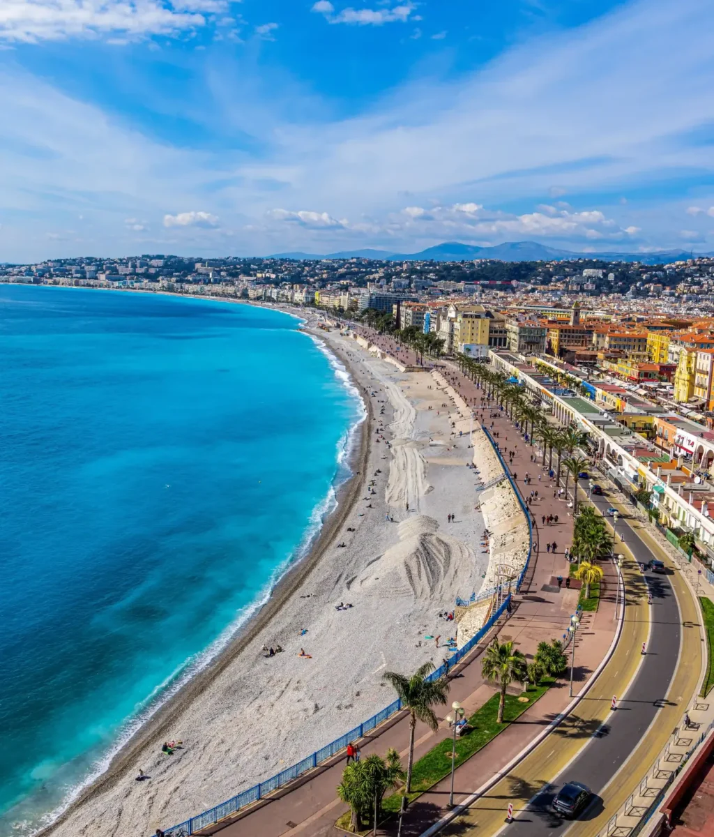 Vue aérienne d'un littoral sinueux aux eaux turquoise, d'une longue plage de galets, de palmiers et de promenades longeant une ville, sous un ciel bleu parsemé de nuages.