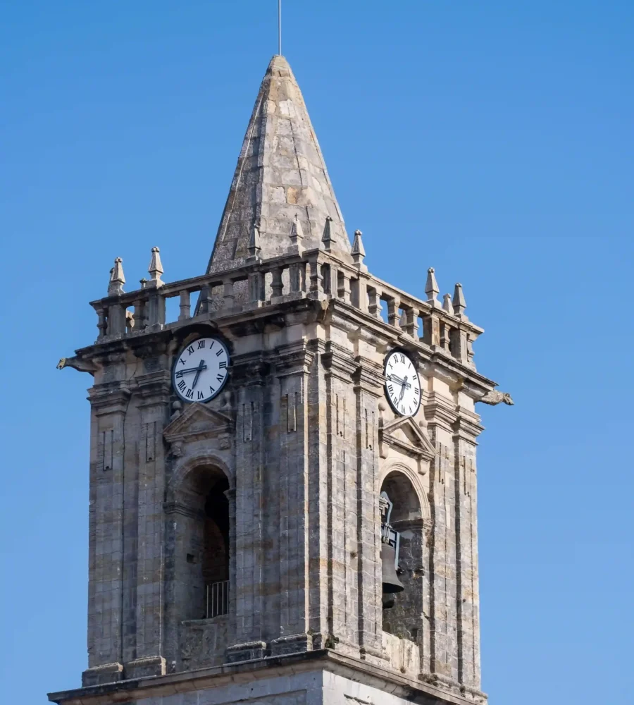 Une tour d'horloge en pierre, dotée de deux grands cadrans, de fenêtres cintrées et d'une flèche pointue, se détache sur un ciel d'un bleu limpide.