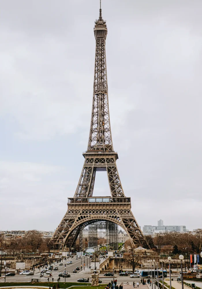 On voit ici la Tour Eiffel à Paris par une journée nuageuse, avec des passants et des voitures circulant dans la rue en contrebas, tandis que des arbres bordent les abords de ce monument emblématique.