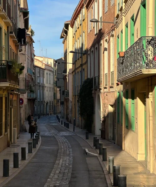 Une ruelle européenne étroite, bordée de bâtiments colorés, de volets verts et de balcons. Deux personnes marchent sur le trottoir sous le soleil matinal, projetant de longues ombres. Des bornes bordent la rue sinueuse pavée.
