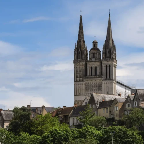 Une grande cathédrale gothique aux deux flèches s'élève au-dessus d'un groupe de maisons et d'arbres verdoyants, sous un ciel bleu partiellement nuageux.