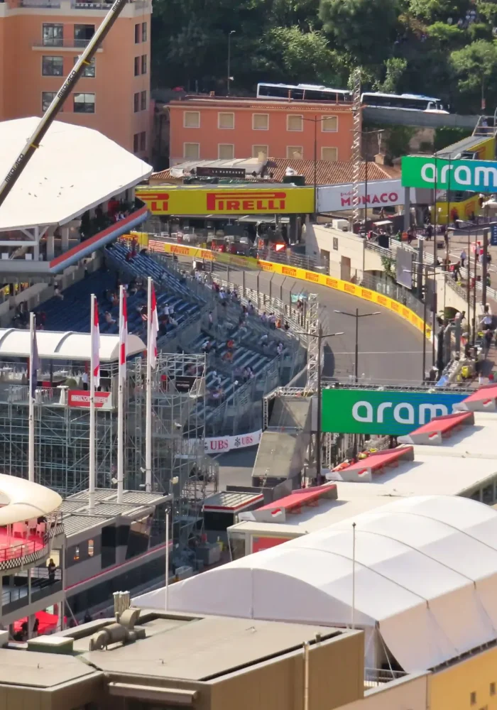 Vue d'un circuit urbain lors des préparatifs du Grand Prix de Monaco, avec des tribunes, des barrières de sécurité, des banderoles et des bâtiments à proximité, sous un soleil radieux.