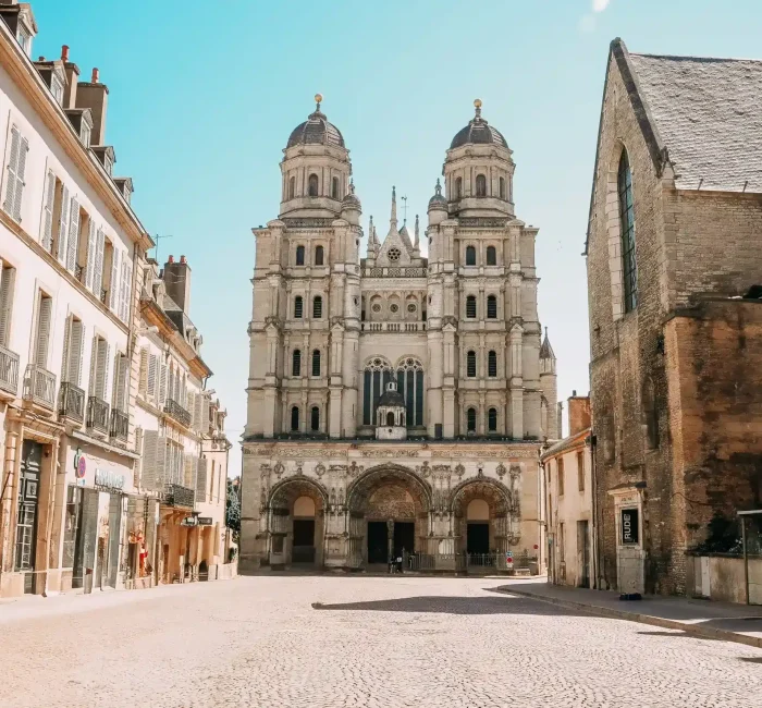 Une vaste place pavée mène à une imposante église historique dotée de deux clochers, entourée de vieux bâtiments en pierre sous un ciel d'un bleu limpide.