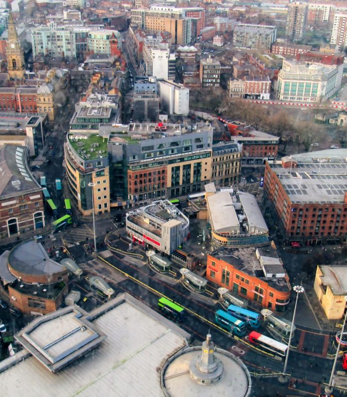 Vue aérienne d'un centre ville animé avec divers bâtiments, rues, bus et voitures. L'architecture comprend à la fois des structures modernes et historiques, et la circulation et les piétons sont visibles dans tout le paysage urbain.